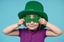 Funny Little Child Girl In Green Patricks Hat With Clover Glasses On Color Background. St. Patricks Day Celebration. Advertising Space