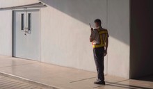 Asian Security Guard In Safety Vest Walking On Sidewalk And Using Walkie Talkie Or Portable Radio Transmitter With Sunlight And Shadow On Surface Of Staff Room Door In Grey Cement Wall Background