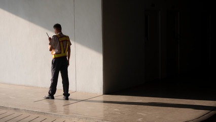  Asian security guard in safety vest walking on sidewalk of parking garage, he using walkie-talkie or portable radio transmitter with sunlight and shadow on surface of gray cement wall background
