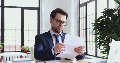 Wall Mural - Confident businessman wearing suit and tie doing paperwork reading paper letter sitting at office desk. Young male ceo executive opening envelope checking postal mail correspondence at workplace.