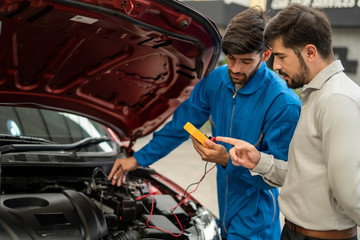 The mechanic in the blue jumpsuit is checking the engine in front of the red open car trunk and the man who is the  car’s owner is talking with the checker who putting the right hand at his forehead