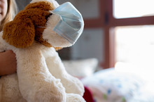 Childhood Fear Of The Virus. Girl Hugging Her Cuddly Toy In Her Room. Cuddly Toy With Mask. Coronavirus Concept