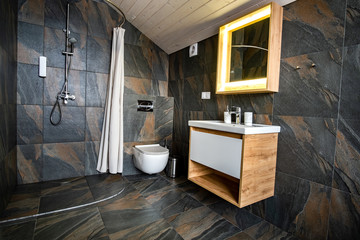 Interior of modern stylish bathroom with black tiled walls, curtain shower place and wooden furniture with wash basin and big illuminated mirror.