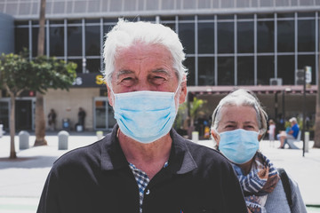 close up of two faces of pensioners and seniors wearing medical mask to prevent virus - airport at the back and safe travelers lifestyle and concept - seriously man and woman looking at the camera