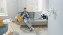 Close Up Object Shot Of A Modern Wi-Fi Surveillance Camera With Two Antennas On A White Wall In A Cozy Apartment. Man Is Sitting On A Sofa In The Background.