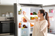 © New Africa - Young woman with apple near open refrigerator in kitchen