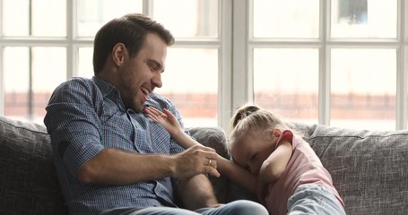 Wall Mural - Happy young father parent tickling small laughing kid daughter. Overjoyed cute little blonde preschool child daughter having fun with joyful daddy, relaxing together on cozy sofa in living room.