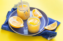 Two Jars Of Homemade Lemon Curd Or Custard And Fresh Lemons On A Blue Ceramic Plate. Yellow Paper Background. Selective Focus.