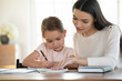 © fizkes - Head shot focused attentive little girl studying, doing homework with mother. Affectionate young mother teaching small daughter reading writing. Female teacher giving private lessons to kid at home.
