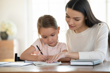 Head shot focused attentive little girl studying, doing homework with mother. Affectionate young mother teaching small daughter reading writing. Female teacher giving private lessons to kid at home.