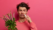 Indoor shot of frustrated man has watery red eyes, runny stuffy nose, uses drops, suffers from hay fever, has allergy on blooming plant, poses on pink background. Spring rhinitis, reaction on trigger
