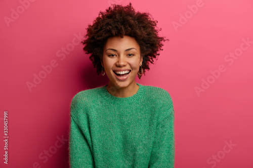 Fotografía Portrait of good looking curly ethnic woman smiles broadly, enjoys day off, has happy talk with interlocutor, discusses holiday preparation, wears green sweater, isolated on pink background