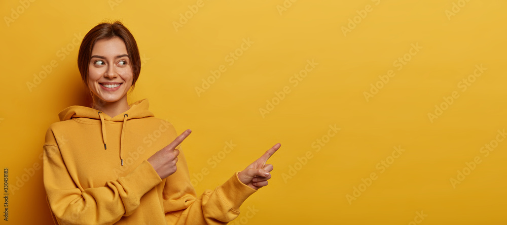 Smiling dark haired woman gives advice and points right, promots product, wears casual sweatshirt, poses against yellow background, indicates at advertisement, introduces promo with pleased expression - obrazy, fototapety, plakaty 