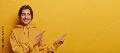 Fotografía Smiling dark haired woman gives advice and points right, promots product, wears
