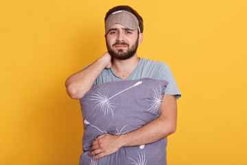 Studio shot of young guy with beard posing isolated over yellow background, wearing gray t shirt, touches his neck, male hugs his pillow, ready for falling asleep, man posing with mask on his forehead