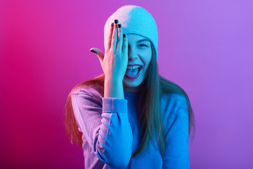 Happy beautiful girl covering one eye, model wearing sweater and knit cap, portrait of woman isolated over pink neon background, lady standing with widely opened mouth. People emotions concept.