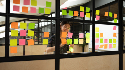 Wall Mural - Excited Asian businesswoman wearing glasses making notes on stickers, smiling leader, executive manager planning project, organizing team work, writing creative ideas on stickers on glass wall