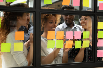 Wall Mural - African American leader with diverse employees team writing creative ideas, tasks on sticky papers on glass scrum board close up, multiethnic colleagues discussing new project plan at meeting