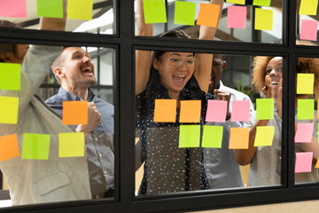 Wall Mural - Asian leader with diverse employees team celebrating business success, achievement at meeting, standing behind glass wall with colorful stickers close up, colleagues excited by finished project