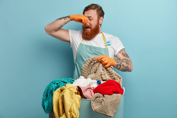 Redhead man covers nose, feels bad smell, disgusting aroma from dirty laundry, going to wash with liquid powder, wears rubber gloves and apron, busy doing housework during weekend. What stench!