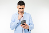 Fototapeta Na ścianę - stylish brunette man in a blue classic jacket looks carefully at a smartphone on a white background