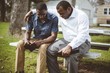African-American male friends sitting on the bench praying with the Bible in their hands