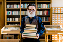 Portrait Of Attractive Bearded Old Man Wearing Shirt And Leather Vest, High School Teacher Or Librarian, Holding Books In Hands, Standing In Vintage Library Interior. Library Worker, Happy Book Day