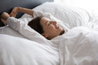 © fizkes - Head shot side view young happy woman lying in bed under duvet, raising arms, stretching back muscles, feeling refreshed in morning. Smiling brunette lady enjoying good holiday time after wake up.