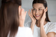 Attractive smiling young brunette woman looking at mirror, touching face. Happy 30s lady satisfied with moisturized soft skin condition after professional cosmetology services, skincare concept.