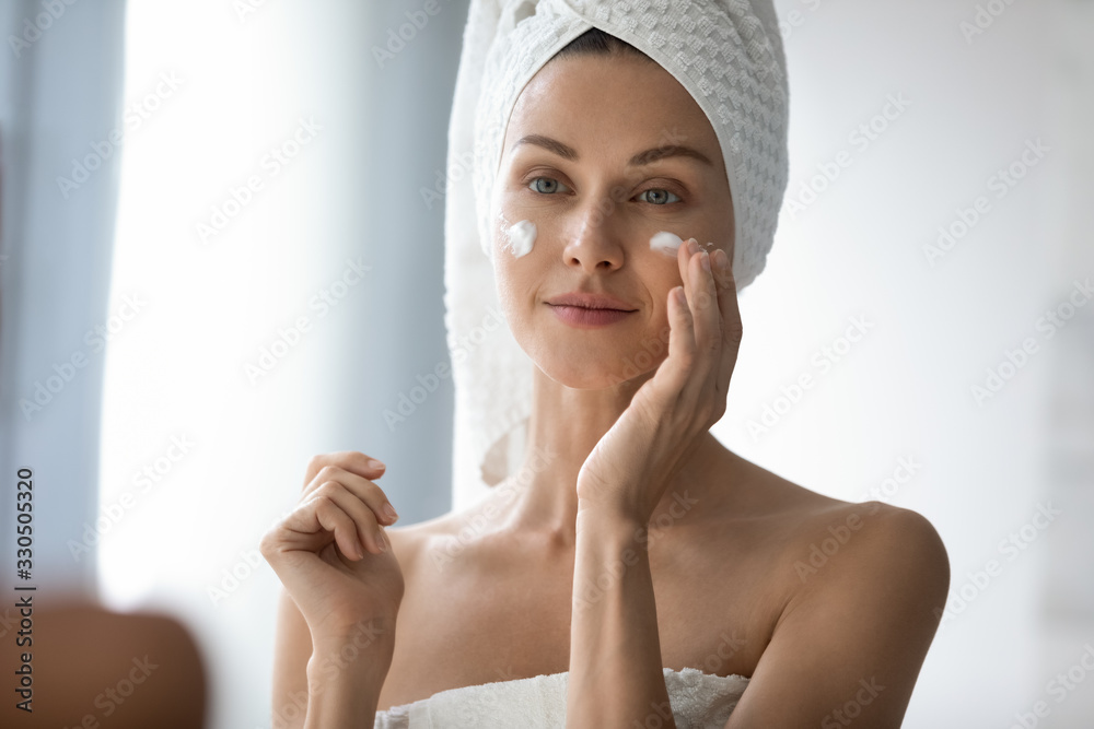Head shot beautiful 30s lady looking in mirror, applying hydrating lotion creme on cheeks, finishing morning domestic skincare routine. Smiling woman grooming herself after showering in bathroom. - obrazy, fototapety, plakaty 