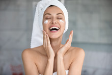 Laughing young woman with wrapped in towel wet hair enjoying applying hydrating creme on face, finishing morning cleansing routine. Joyful lady using professional skincare product alone in bathroom.