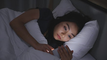 Asian Young Woman Lying On Bed Play Smartphones During Night Time In Bedroom That Turns Off Lights. Read COVID-19 On Mobile Phone. Using P Hone In Low Light Has Health Effect On Eyes. Insomnia Concept