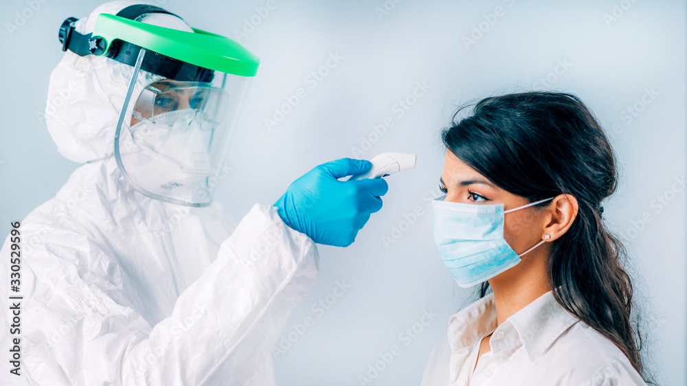 Coronavirus Screening. Medical worker in protective suit checking body temperature with contactless body thermometer, young woman wearing protective mask - obrazy, fototapety, plakaty 