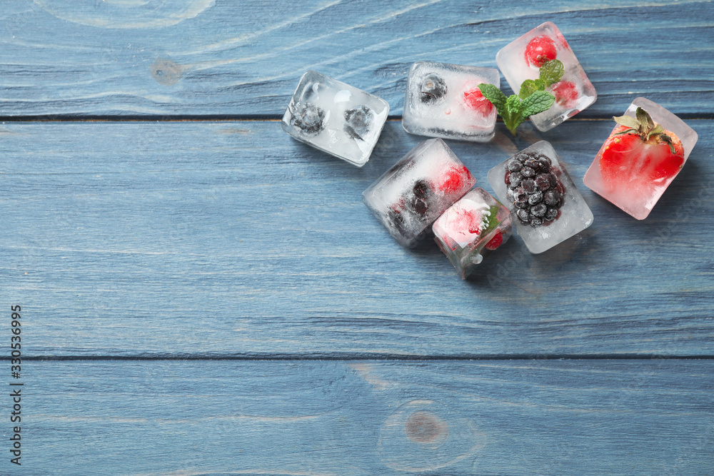 Ice cubes with different berries and mint on blue wooden table, flat lay. Space for text - obrazy, fototapety, plakaty 