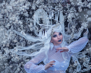 Very close portrait of amazing cute young woman in fairy tale image with long white hair and unusual magic hat on her head in lace dress and dark beautiful makeup surrounded by white flowers