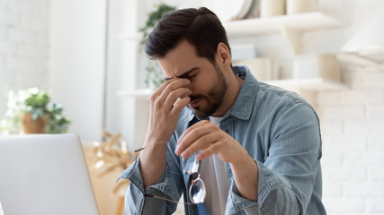 Tired unhealthy man taking of glasses, massaging nose bridge, feeling unwell, suffering from eye strain after long laptop use, exhausted young male student, freelancer relieving dry eyes syndrome