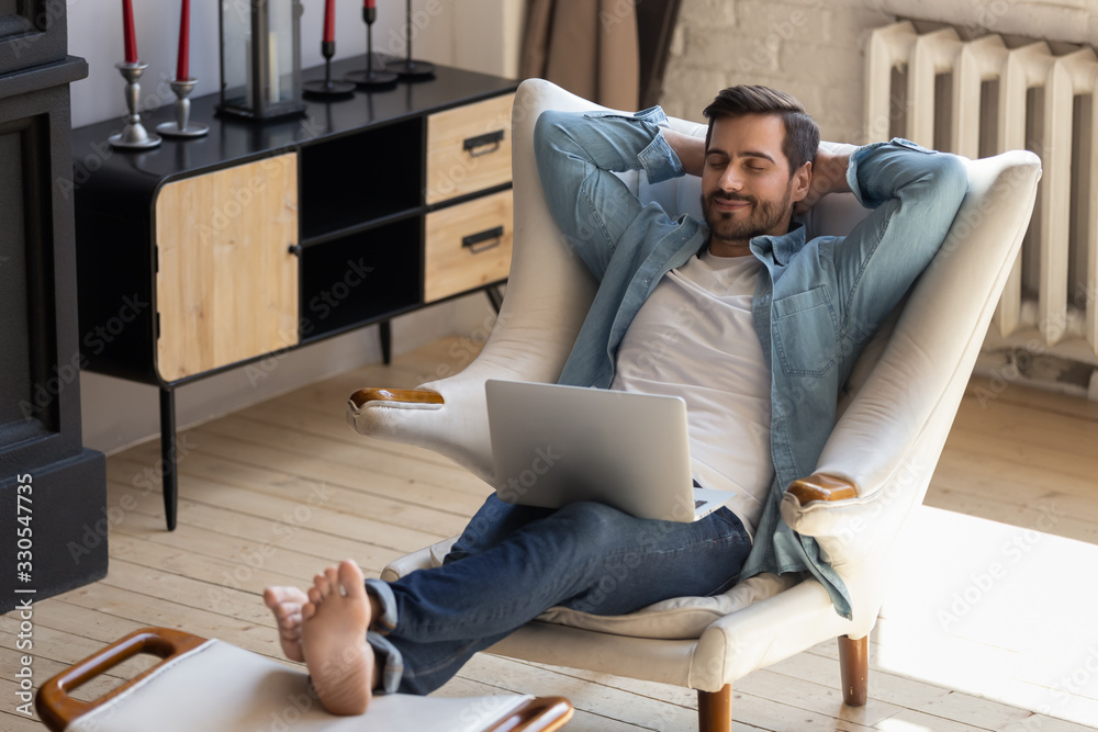 Calm young man relaxing with laptop on cozy armchair, peaceful male with closed eyes and hands behind head distracted from online work, enjoying lazy leisure time, daydreaming during break at home - obrazy, fototapety, plakaty 