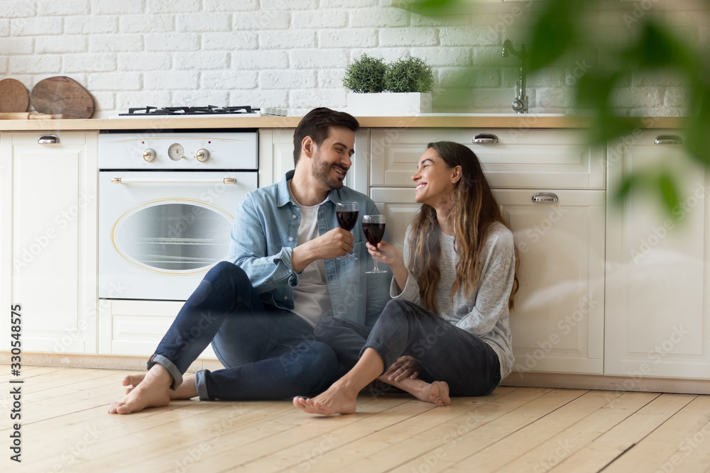 Happy loving couple clinking red wine glasses in modern kitchen, sitting on warm wooden floor together, smiling wife and husband celebrating relocation or anniversary, having fun, romantic date - obrazy, fototapety, plakaty 