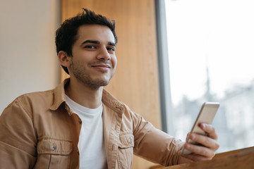 Happy asian guy holding cellphone, chatting, booking tickets, looking at camera and smiling. Handsome Indian man using smart phone with mobile app for ordering food online