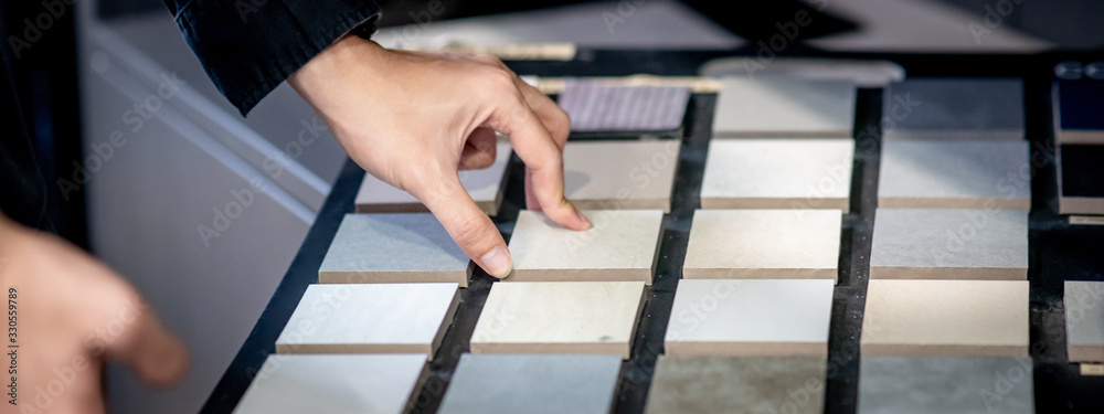 Male architect or interior designer hand choosing ceramic texture sample from swatch board in design studio. Floor and wall finishing material for architecture and construction industry. - obrazy, fototapety, plakaty 
