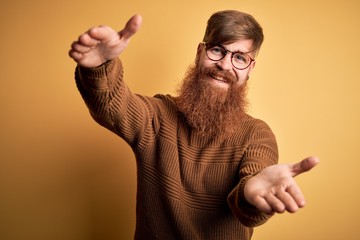 Poster - Handsome Irish redhead man with beard wearing glasses and winter sweater over yellow background looking at the camera smiling with open arms for hug. Cheerful expression embracing happiness.