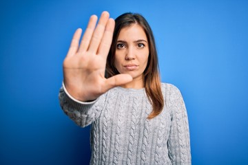 Sticker - Beautiful young woman wearing casual wool sweater standing over blue isolated background doing stop sing with palm of the hand. Warning expression with negative and serious gesture on the face.