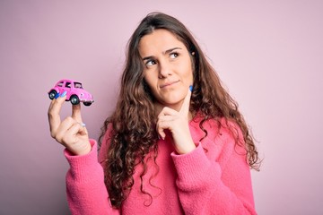 Sticker - Young beautiful woman with curly hair holding small car over isolated pink background serious face thinking about question, very confused idea