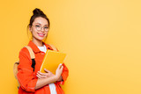 Fototapeta  - cheerful student in glasses looking at camera while holding notebook isolated on yellow