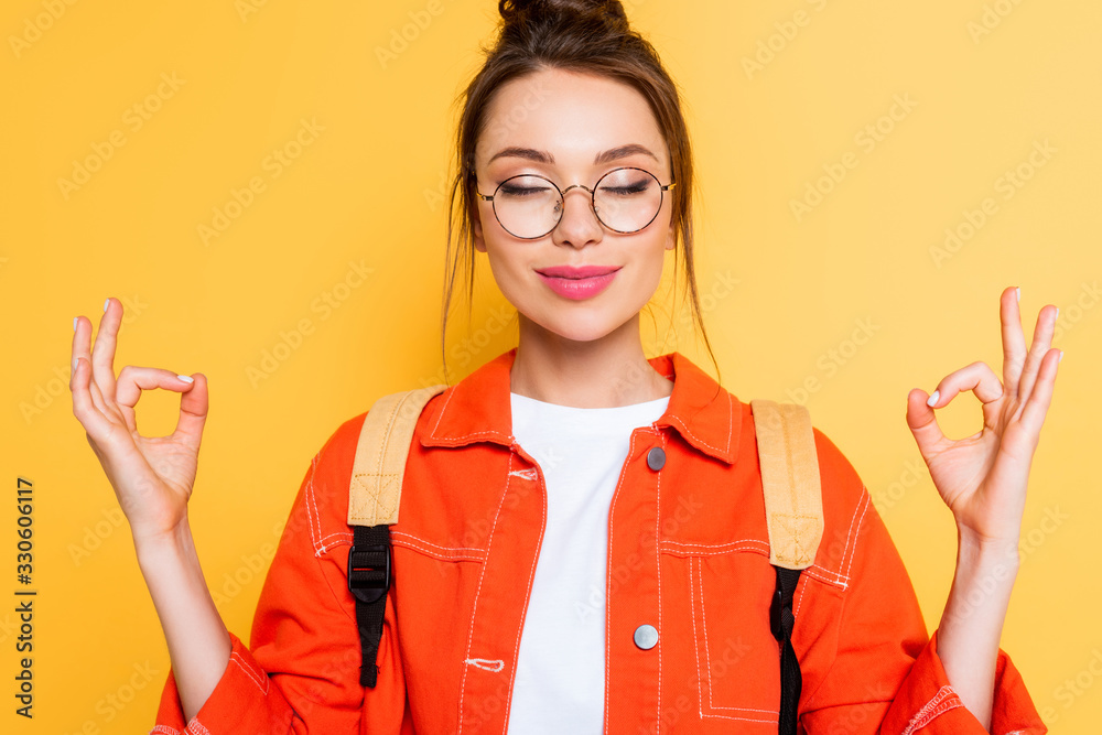 smiling student in eyeglasses standing in meditation pose with closed eyes isolated on yellow - obrazy, fototapety, plakaty 