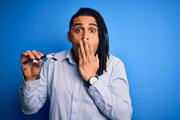 Young african american afro man with dreadlocks holding glasses over blue background cover mouth with hand shocked with shame for mistake, expression of fear, scared in silence, secret concept