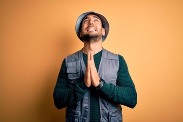 Sticker - Handsome tourist man with beard on vacation wearing explorer hat over yellow background begging and praying with hands together with hope expression on face very emotional and worried. Begging.