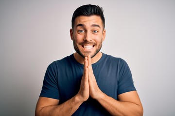 Canvas Print - Young handsome man wearing casual t-shirt standing over isolated white background praying with hands together asking for forgiveness smiling confident.