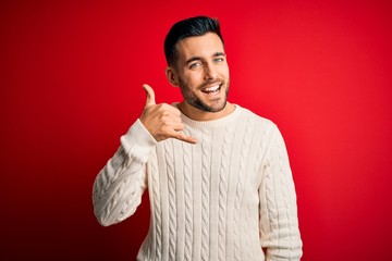 Young handsome man wearing casual white sweater standing over isolated red background smiling doing phone gesture with hand and fingers like talking on the telephone. Communicating concepts.