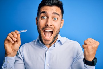 Wall Mural - Young handsome man holding dental aligner tooth correction over blue background screaming proud and celebrating victory and success very excited, cheering emotion